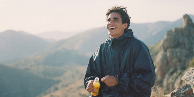 accidentes-express hombre joven sonriente con chubasquero oscuro de pie en la cima de una montaña rocosa. sostiene una botella naranja.