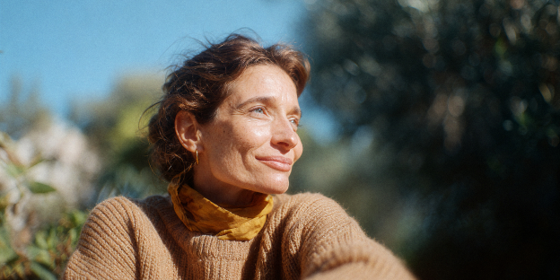 accidentes-mujer.webp mujer adulta sonriendo con jersey de punto marrón y pañuelo, luz solar brillante, follaje verde y cielo azul.