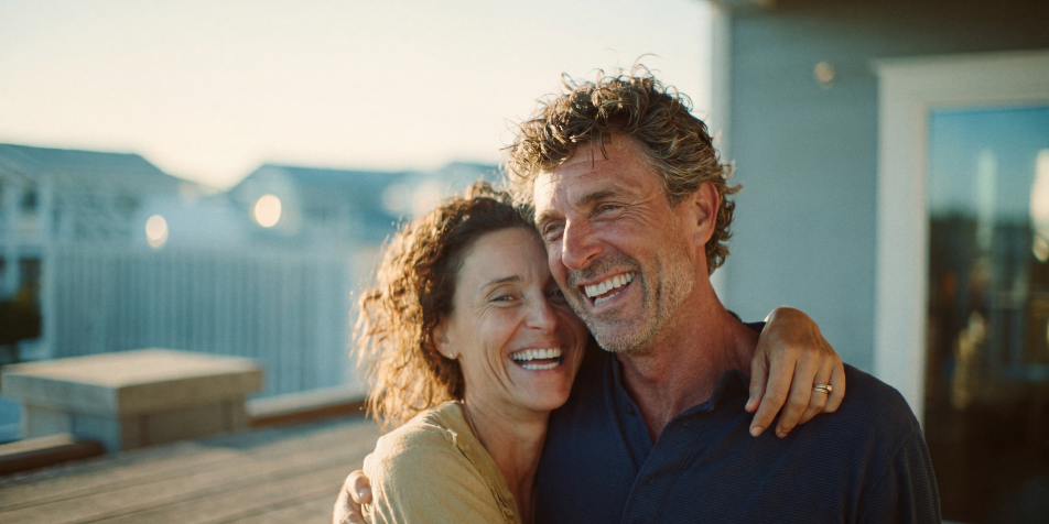 Ahorro Inversion - Pareja sonriente abrazada dos adultos de piel clara se abrazan y ríen al aire libre con un fondo de casas y luz cálida.