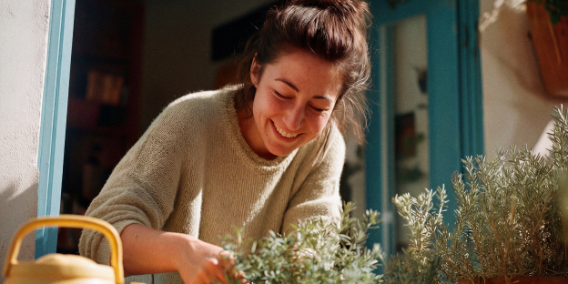 ahorro_garantizado_flexible una mujer sonriente atiende plantas verdes en macetas; un marco de ventana azul y una regadera amarilla aparecen