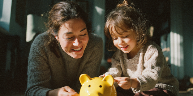 ahorro_garantizado_infantil una mujer y una niña sonriendo miran una hucha amarilla de cerdo en el suelo, la niña introduce un objeto.