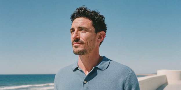 un hombre de pelo oscuro y barba en primer plano, viste un polo azul claro, mirando hacia el cielo azul y el mar bajo un sol brillante.