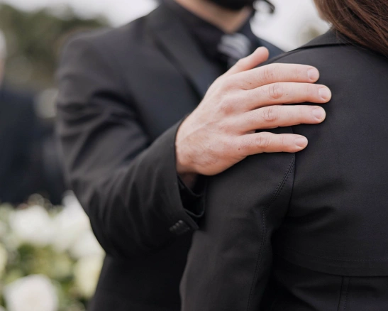 un hombre vestido de negro apoya su mano sobre la espalda de una mujer en un funeral.