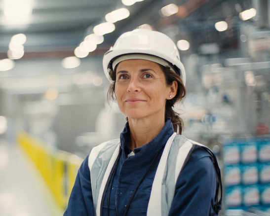 mujer de mediana edad con casco de seguridad blanco y chaleco reflectante azul en interior de nave industrial.