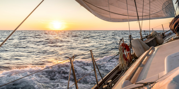 embarcaciones_recreo cubierta de velero navegando en mar con oleaje al atardecer. se ve una vela blanca, un aro salvavidas naranja y el sol bajo sobre el horizonte.