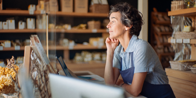 empresas una mujer con delantal azul se reclina en un mostrador de madera con un monitor. detrás hay estanterías llenas de productos.