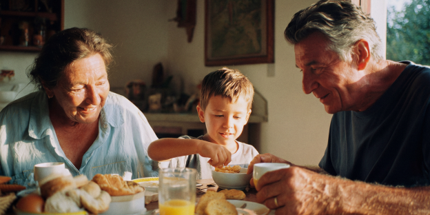 hogar_senior una mujer, un niño y un hombre sentados en una mesa, desayunando con tazas, cereal, pan y zumo.