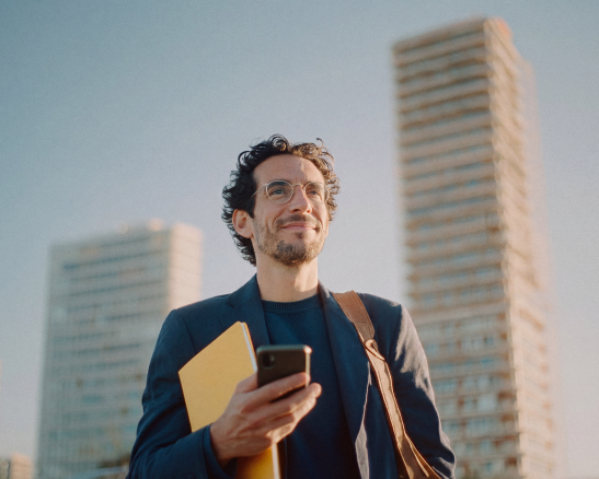 hombre-movil-edificios.webp hombre sonriendo con gafas y chaqueta oscura. sujeta un móvil y una carpeta amarilla. dos edificios altos en el fondo.