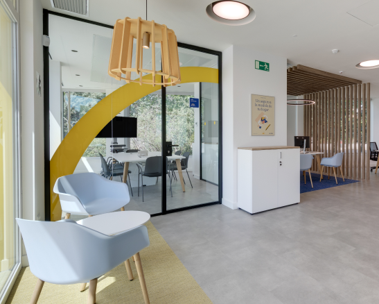 sala de espera de oficina moderna con dos sillones azules, mesa lateral, mampara de cristal y mueble de almacenamiento blanco.