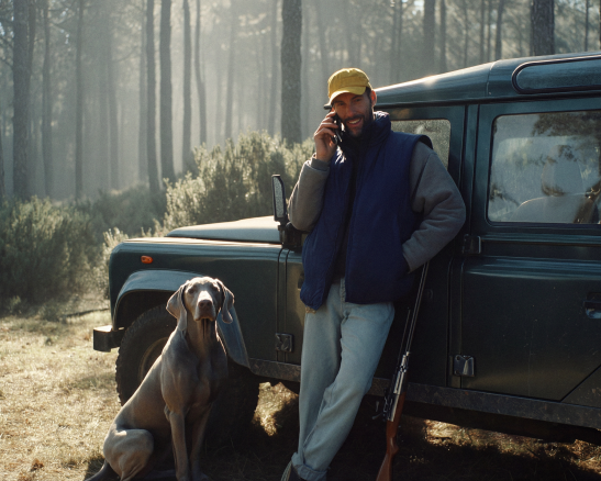 un hombre con gorra y chaleco azul habla por teléfono junto a un perro de raza gris, apoyado en un todoterreno con un rifle en un bosque.