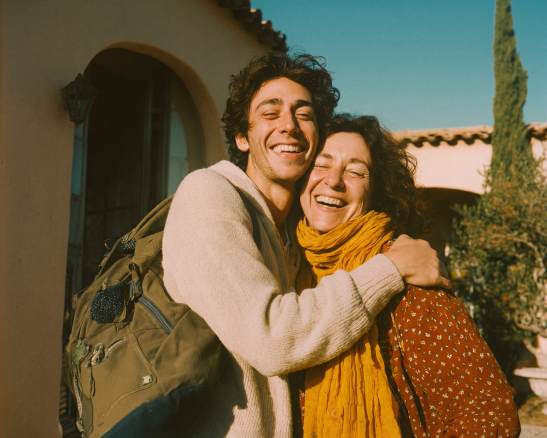 hombre joven con suéter claro y mochila verde abraza a mujer adulta con pañuelo amarillo y camisa estampada frente a una pared beige.