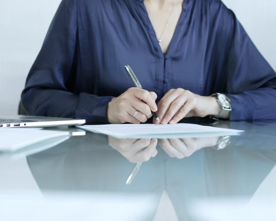 Mujer con blusa azul marino firmando un documento sobre una mesa de cristal, junto a un portátil.