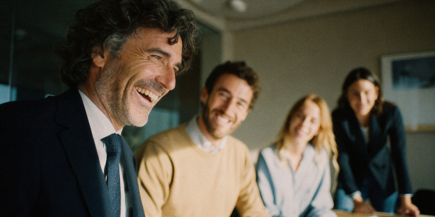 cuatro personas sonriendo en una reunión de oficina. un hombre con traje en primer plano, tres personas sentadas detrás.