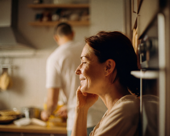 mujer sonriendo sentada en una cocina con gabinetes de madera, un hombre de espaldas está desenfocado en el fondo.